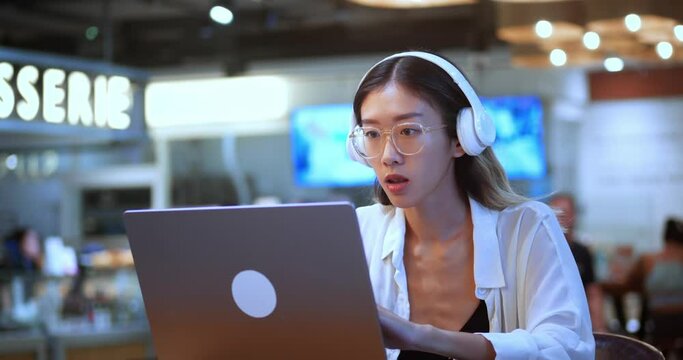 Asian female business person wears headset conference video calling looking at laptop computer sitting at cafe. Young businesswoman manager speaking to webcam communicating in distance remote chat.