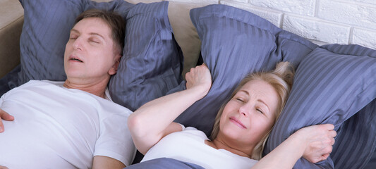 Snoring man. Couple in bed, man snoring and woman can not sleep, covering ears with pillow for snore noise. Middle age couple in bed at home.