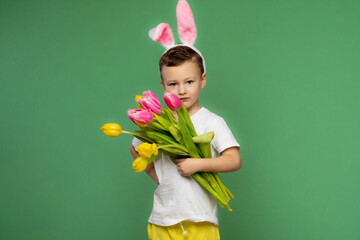 Happy beautiful boy on her head with rabbit ears, with a bouquet of tulips on a green background. Symbol of Easter and spring.