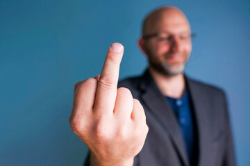 Hand in middle finger gesture in focus, Bald man in business suit out of focus, blue background....