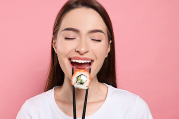 Beautiful young woman eating sushi roll with chopsticks on pink background