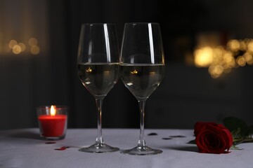 Glasses of white wine, burning candle and rose flower on grey table against blurred lights. Romantic atmosphere