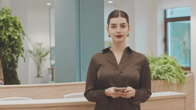 Medium portrait of young beautiful Middle Eastern woman using smartphone and smiling at camera standing ay modern coworking space