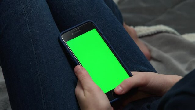 Close up video of cropped male hands using blue smartphone with green screen on knees. PDA, technology and education online, reading articles, surfing internet. High angle view, above shoulder