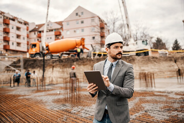 A site chief is scrolling on tablet and visiting works on construction site. There are concrete mixer and workers in a blurry background.