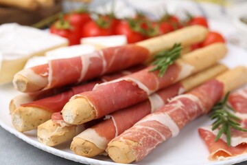 Plate of delicious grissini sticks with prosciutto, cheese and tomatoes on light grey table, closeup