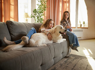 Beautiful purebreed dog, sand color American retriver lying on sofa with two kids, little girls in casual style clothes. Concept of happiness, family, care, animal and vet