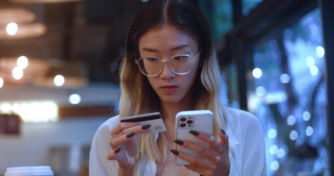 young woman customer holding credit card and smart phone sitting at coffee shop. Asian female teenager shopper using smart phone and credit card for mobile payments making purchase in online store