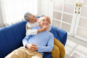 Obraz premium Portrait of happy Asian senior couple living together, hug, touching and embracing with a smile on the sofa in the living room. Retirement living together at home.