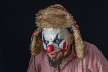 A bearded man wearing a creepy clown mask and a hat with ears looks into the camera as he stands in...