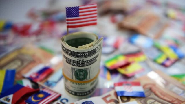 A roll of US dollars with the American flag on top of a other currencies and country flags. Dollar hegemony concept.