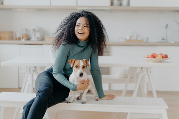 Joyful Afro woman sits at white bench together with dog against kitchen interior, table with plate...