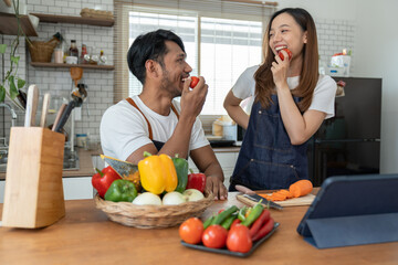 Young Asian couple wearing apron learning how to choose ingredients such as fruits and vegetables to prepare their morning meal via tablet, online. Lifestyle and healthy cooking concept.