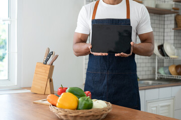 Asian athletic young man in apron using tablet to learn how to cook Choosing ingredients online with internet in happy and relaxed mood inside home kitchen, part-time work concept healthy food.