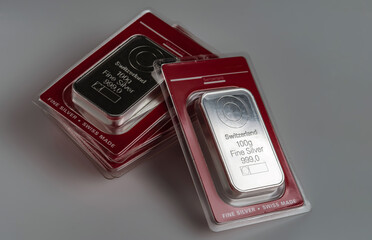 Several minted silver bars weighing 100 grams in transparent blister pack on a grey background.