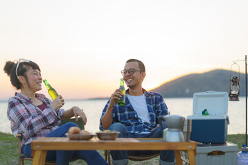 Asian couple drinking beer from bottle in their camping area with lake in the background during sunset.
