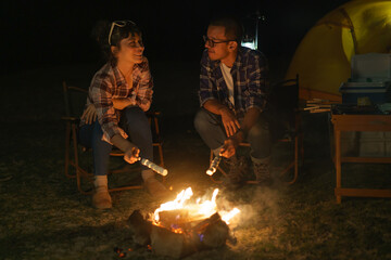 Asian couple roasting marshmallows together at a campfire where they set up a tent to camp by the...