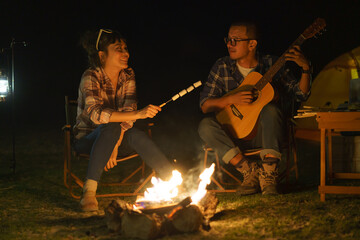 Asian woman toasting marshmallows while her boyfriend plays guitar At the campfire where they set...