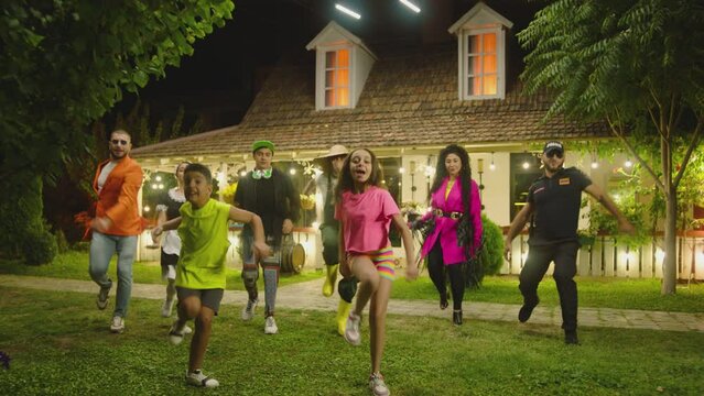 Group of funny , stylish multi aged dancers dancing near village house at night . Kids dancing with house workers . Multi ethnic people having fun . Beautiful background with different lights .