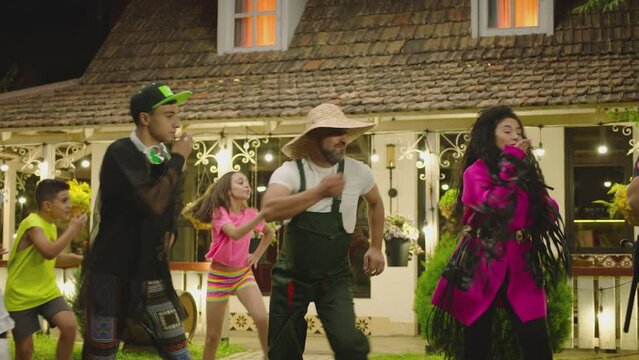 Group of funny , stylish multi aged dancers dancing near village house at night . Kids dancing with house workers . Multi ethnic people having fun . Beautiful background with different lights .