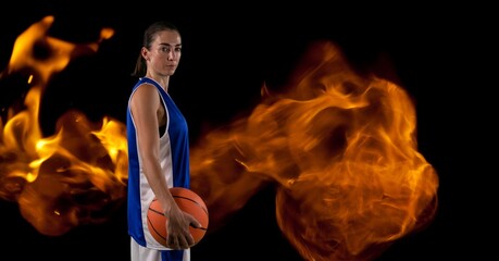 Composition of female basketball player standing holding ball over flames on black background
