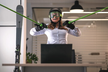 Woman in an office with ski helmet and ski goggles on the head holds ski poles and mimics skiing in front of laptop