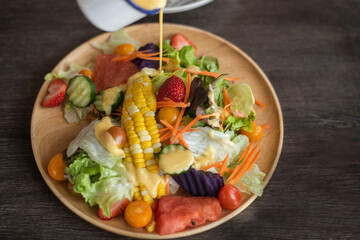 Vegetable and fruit salad in light brown wooden plate On top is pouring dressing. May be slightly blurred by movement The salad plate is placed on a dark brown wooden table