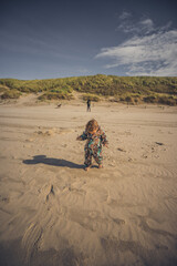 Kleinkind läuft am Stand durch den Sand und hat Spaß, im Hintergrund beobachtet Mutter mit Hund...