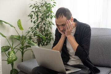 Confused man sitting on couch with computer on lap in living room, upset frustrated male reading...