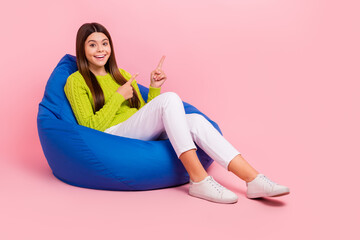 Full length photo of cute young girl sitting bean bag excited point empty space wear trendy green outfit isolated on pink color background