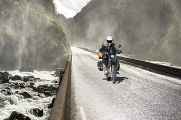 Motorbike driving fast on wet canyon valley road and bridge across river. Motobiker journey in Norway. - 572249259