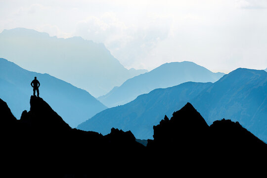 One Man reaching summit after climbing and hiking enjoying freedom and looking towards mountains silhouettes panorama during sunrise.