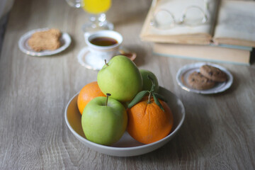 Cup of tea, plates with cookies, glass of orange juice, books, reading glasses, bowl of fruit and candles on the table. Selective focus.