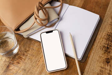 Top view of the cellphone with a white screen with pencil, notepads, and fashion bag on a desk.