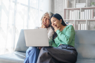 60s asian mother elderly sitting on sofa with young asia female daughter together in living room. watch movies series online or shopping, using laptop computer