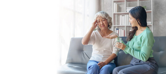 60s asian mother elderly sitting on sofa with young asia female daughter together in living room. give medicine and water