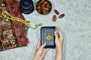 lantern and Holy Quran with Arabic calligraphy meaning of Al Quran and prayer mat , rosary muslim and bowl dates fruits and woman hands holding a holy book