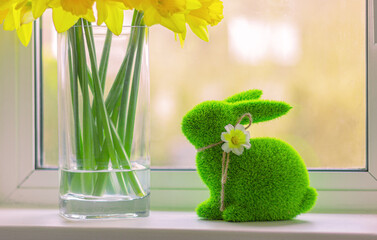green Easter rabbit made of artificial grass and yellow flowers in vase. Spring bunny figurine and daffodils