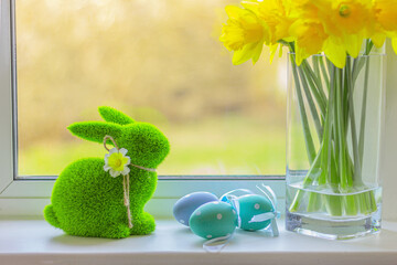 Easter rabbit on windowsill with colorful eggs and spring flowers. green grass bunny figurine.