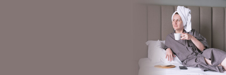 Middle-aged woman with towel on her head drinks coffee in bed. Woman is resting in hotel and relaxing. Сoncept of taking care of yourself, have a nice morning. Banner with free space for text