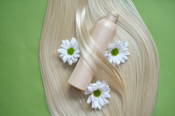 Fototapeta na wymiar eco-friendly hair care concept. ply of blonde hair with white daisies and a beige shampoo bottle lying on a green background.