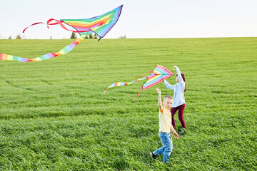 Smiling girl and brother boy running with flying colorful kites on the high grass meadow. Happy childhood moments