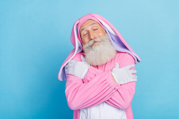 Portrait of cheerful positive man dressed pink rabbit costume cuddle himself enjoy comfort sleepwear isolated on blue color background