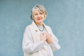 Outdoor portrait of beautiful 55 - 60 year old woman posing next to blue background wall, holding hands next to chest, thank you sign - 572274223
