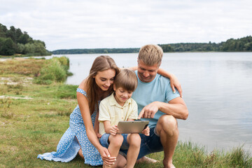 A happy family sit on the bank of a picturesque river and play on the tablet together. Mom is wearing a blue dress, dad and son are in denim shorts and t-shirts.
