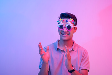 A young male celebrant clapping or giving applause. Wearing hilarious birthday novelty shades and having a good time. Lit with blue and pink neon colors.