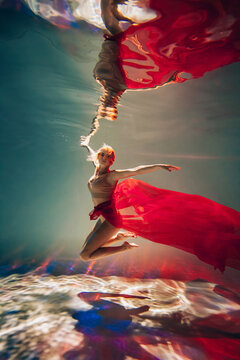 Art work. A slender, tanned girl with an athletic figure and blond hair, with red material and light underwear, in a ballet pose underwater in the pool. Aesthetic image for your design or decoration.