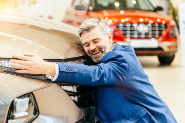 Joyful man, car dealership customer hugging his freshly bought car in showroom.