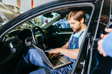 Experienced bearded expert male specialist in blue overall sitting at car with laptop at car service. Repair, maintenance and people concept.