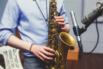 Fototapeta premium View of a saxophone player in headphones during rehearsal, recording sound for new album song at studio, saxophonist musician in front of microphone with musical band orchestra, music production